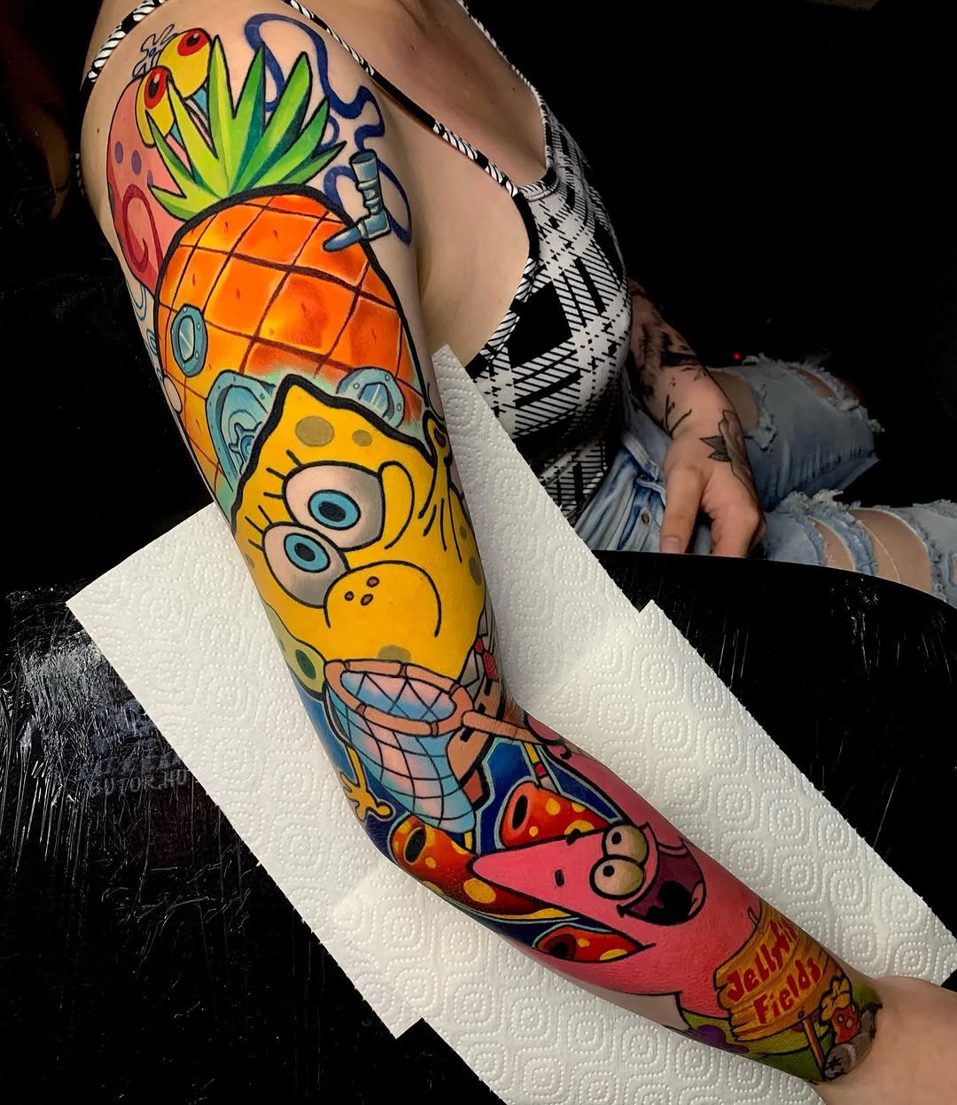 Colorful leg sleeve tattoo featuring a yellow character with large eyes, pineapple, strawberries, and red creature with text, displayed on white paper.