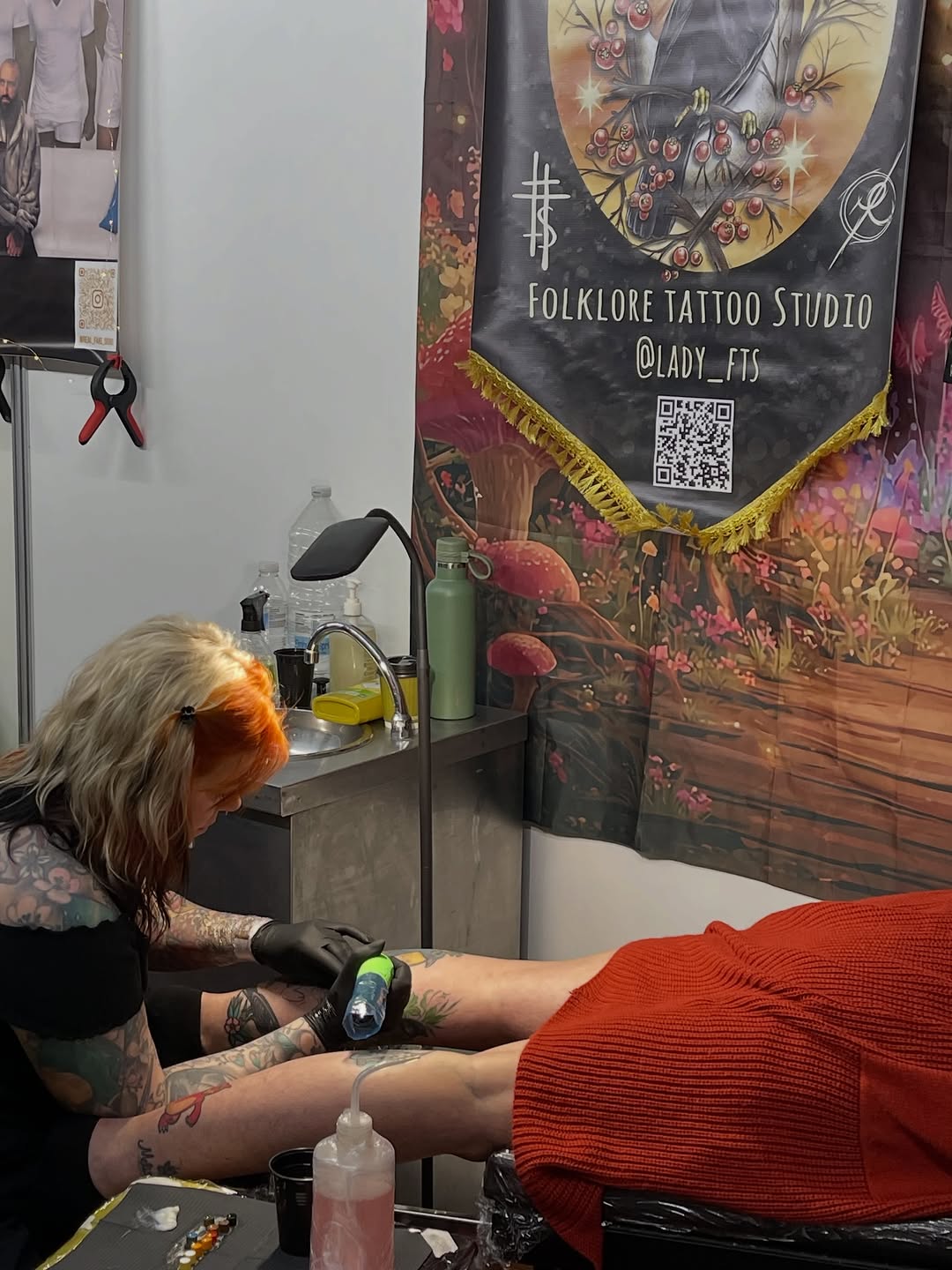 Tattoo artist with orange-tinted hair applies a colorful design to a client's leg wearing a red sweater at Folklore Tattoo Studio.