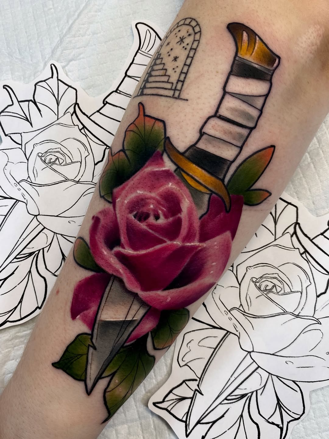 Colorful rose tattoo with green leaves on an arm, featuring two decorative brushstroke elements above the flower in gold and black.