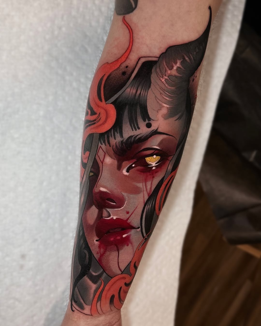 Demon-faced character with horns, golden eye, and red facial features tattooed on forearm in red, black, and skin tones.