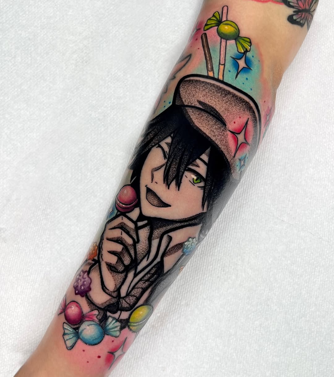 Colorful arm tattoo featuring an anime-style character with black hair and green eyes, surrounded by lollipops, paintbrushes, and candy elements in pink, blue, and yellow tones.