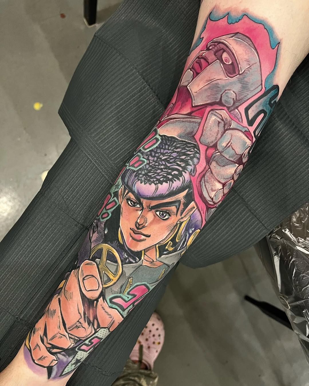 Colorful sleeve tattoo featuring two characters with pink and teal backgrounds, detailed facial features, and gold accessories on the forearm.