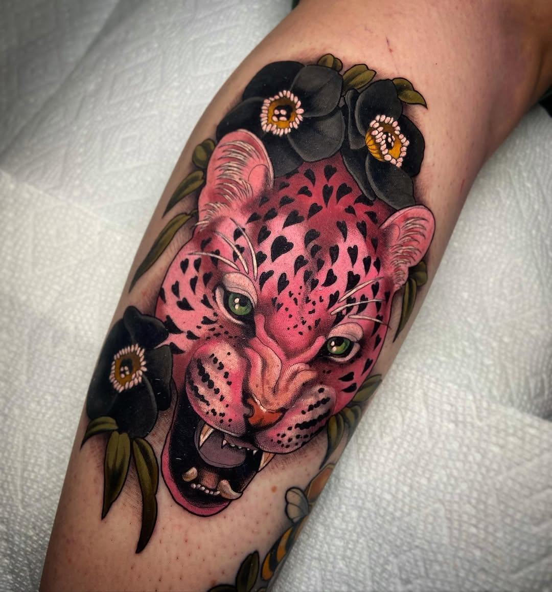 Pink leopard head with green eyes surrounded by black flowers with golden centers on the forearm.