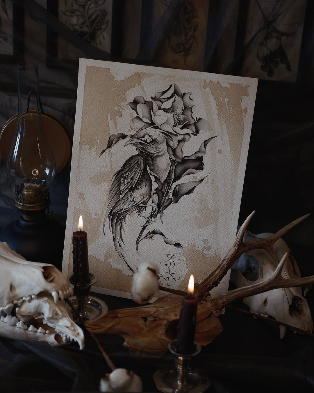 Detailed black ink bird with rose flower and botanical elements displayed on cream paper, surrounded by candles, antlers, and skulls in dark aesthetic setting.