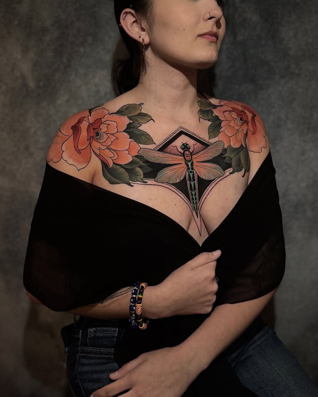 Woman displaying a chest tattoo featuring coral peonies on the shoulders and a detailed dragonfly centered on the upper chest, in orange, green, and black ink.