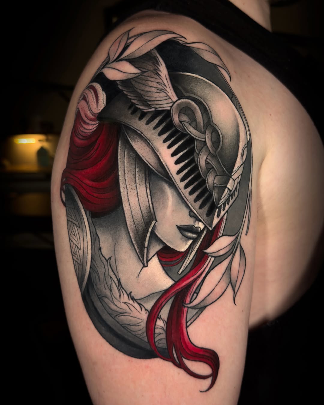 Woman's upper arm tattoo featuring a winged helmeted figure with red fabric accents, grey and black shading, and decorative leaf elements.