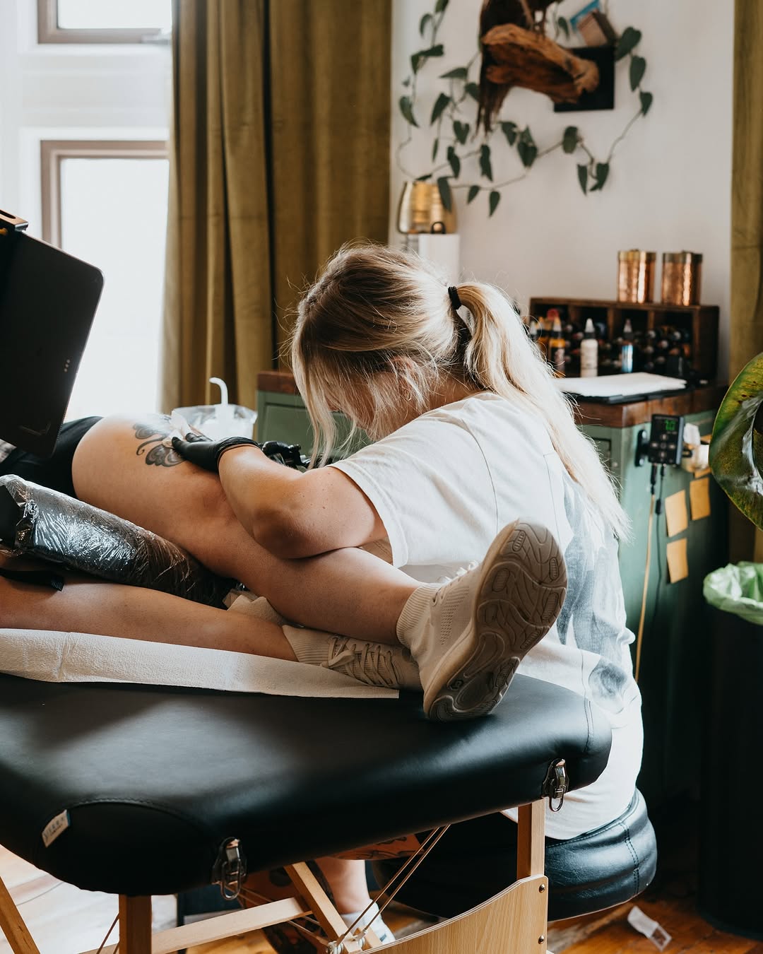 Tattoo artist applying a brown wing design on a client's upper arm in a studio setting with green curtains and wooden furnishings.