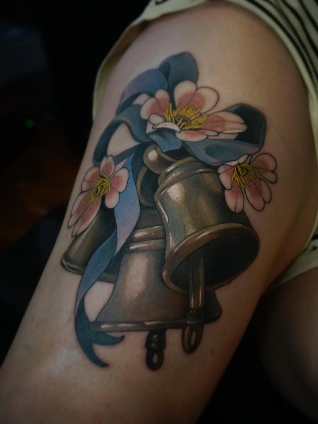 Watering can with pink and blue flowers featuring yellow stamens and blue ribbon, tattooed on upper arm in color.