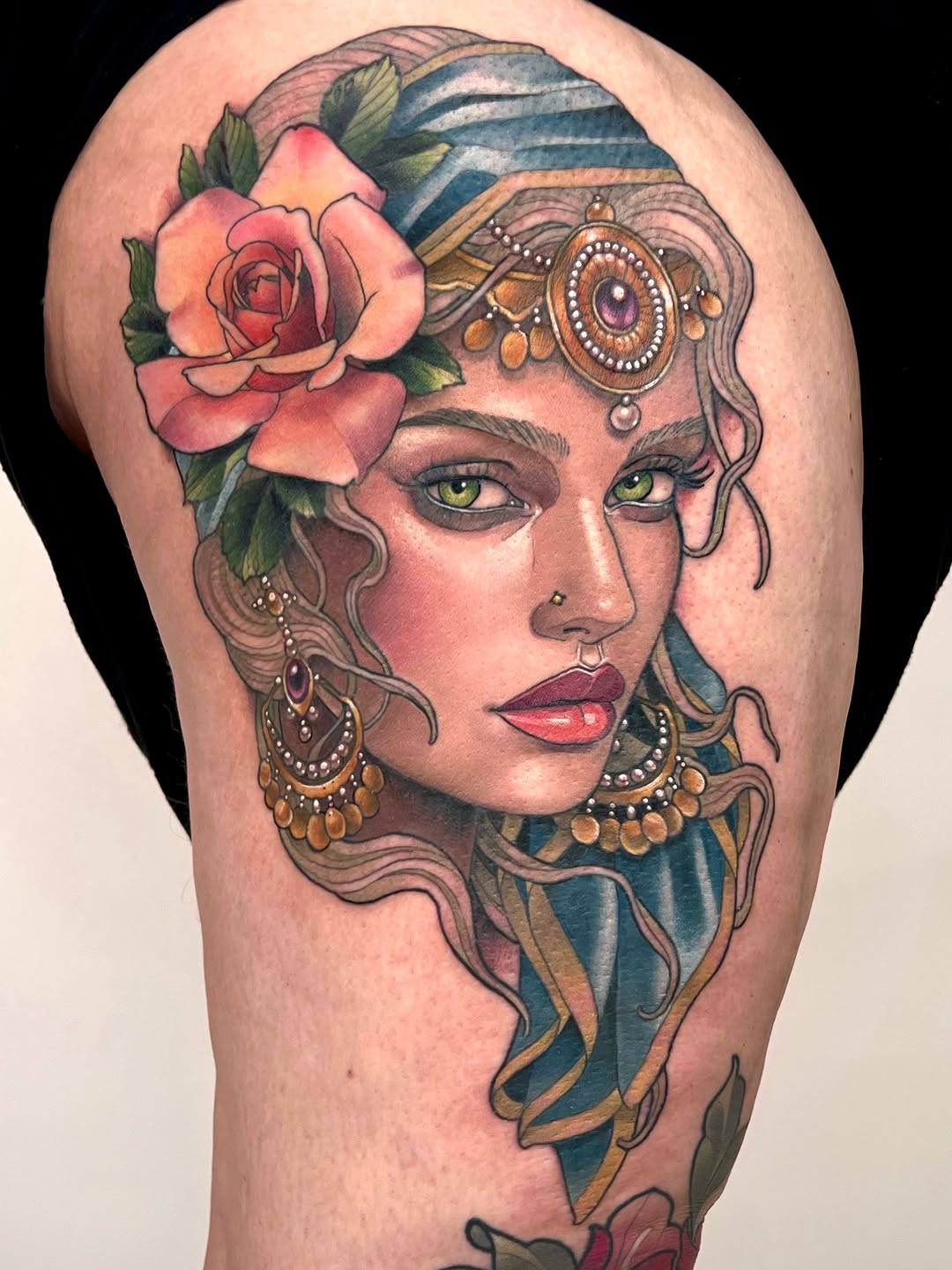 Colorful shoulder tattoo of a woman's face with green eyes, coral rose, ornate eye patch, gold jewelry, and flowing blue and teal hair ribbons.