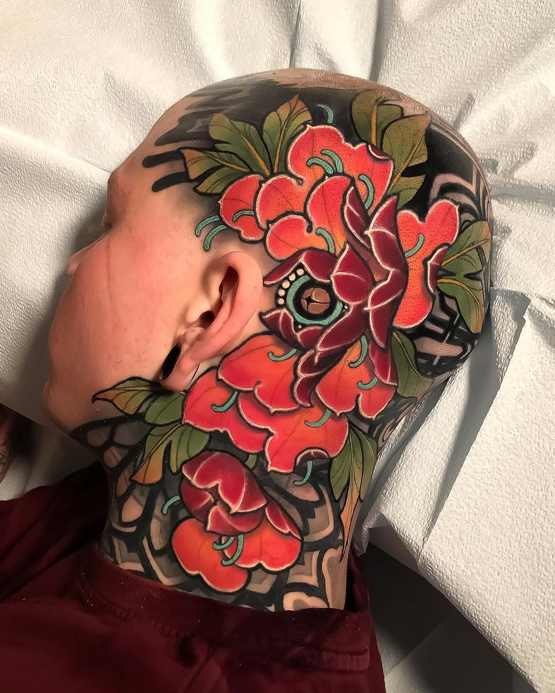 Profile view of a head and neck tattoo featuring large red flowers with green leaves and dark outlines on a skin-tone background.
