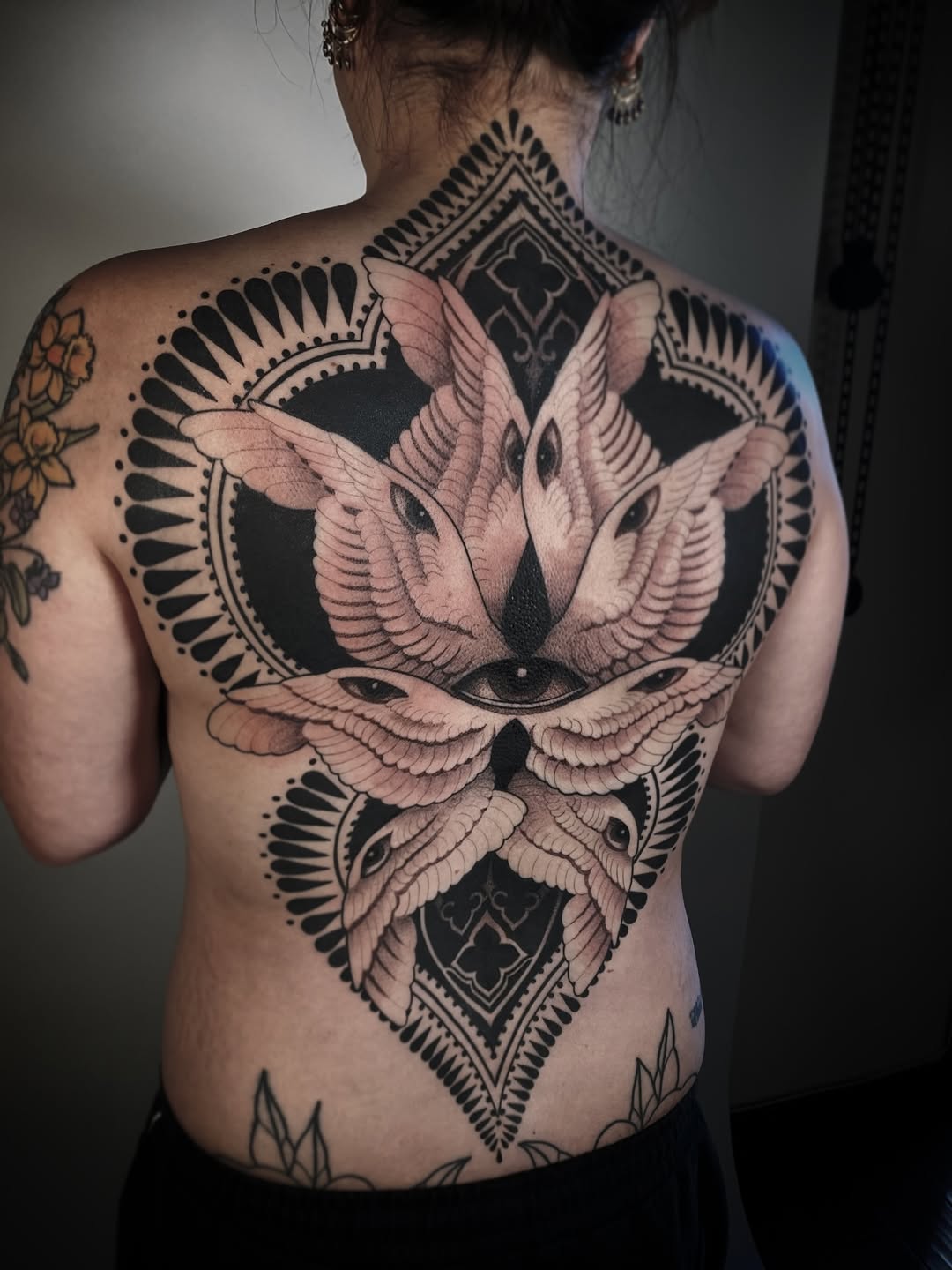 Back tattoo featuring three winged birds surrounding geometric diamond shapes with decorative fan patterns in black and pink ink.