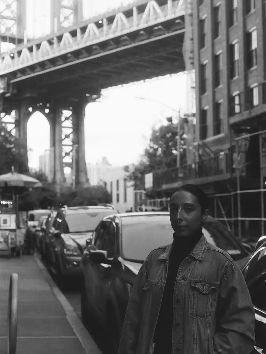 Black and white portrait photograph of a person wearing a denim jacket standing on an urban street beneath an elevated bridge structure.