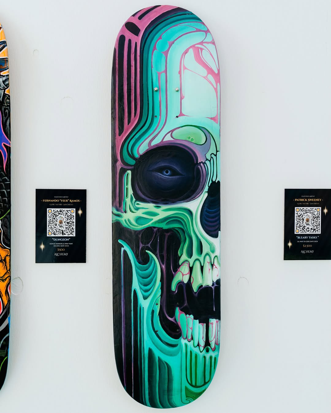 Skateboard deck featuring a neon green and pink skull with an eye design, displayed on a white gallery wall with an artist plaque.