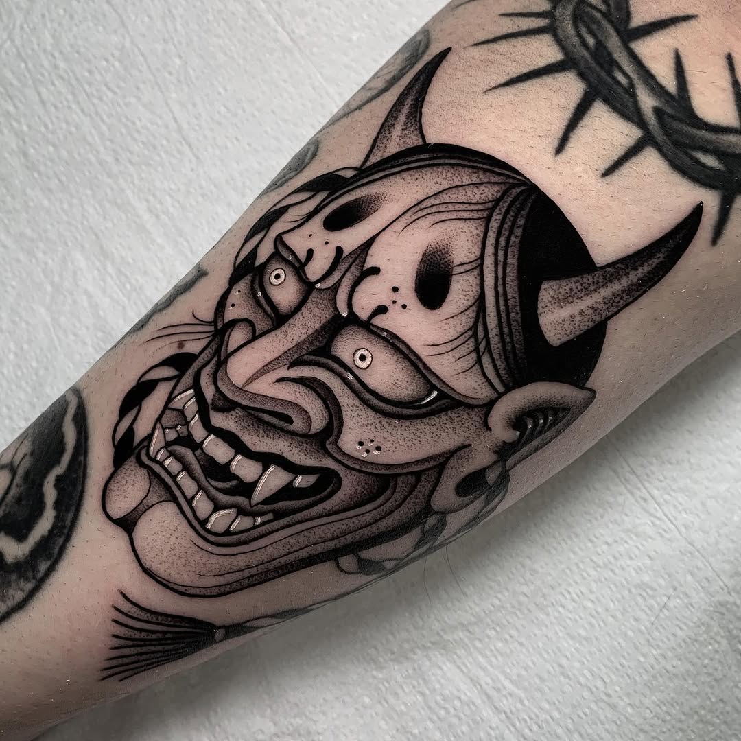 Demon or oni mask tattoo with intricate linework and shading on a forearm, featuring expressive eyes and detailed facial features in black ink.
