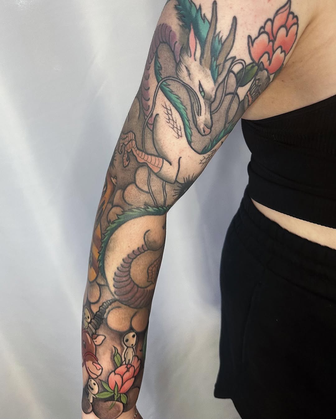 Full-color arm sleeve tattoo featuring anatomical heart with flowers, botanical elements, and small skeletal figures in coral, green, and tan tones.