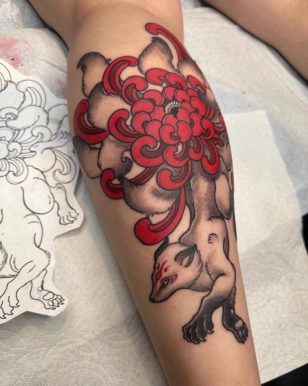 Tattoo on forearm depicting a mythical creature with elaborate red scrollwork features, shaded animal heads, and detailed linework in red and black ink.