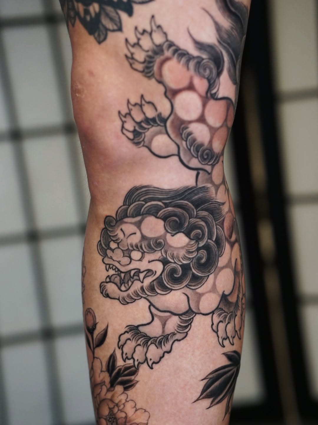 Lion head tattoo on leg with clouds and foliage, rendered in black ink with peach and tan shading throughout the design.