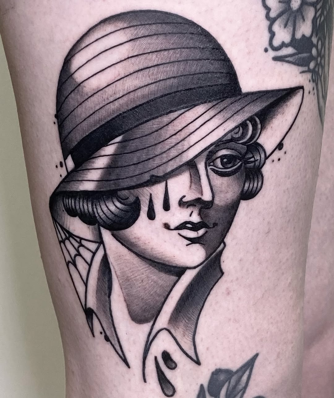 Portrait tattoo of a woman wearing a wide-brimmed hat with decorative curled hair and spider web detail on neck, rendered in black ink on upper leg.