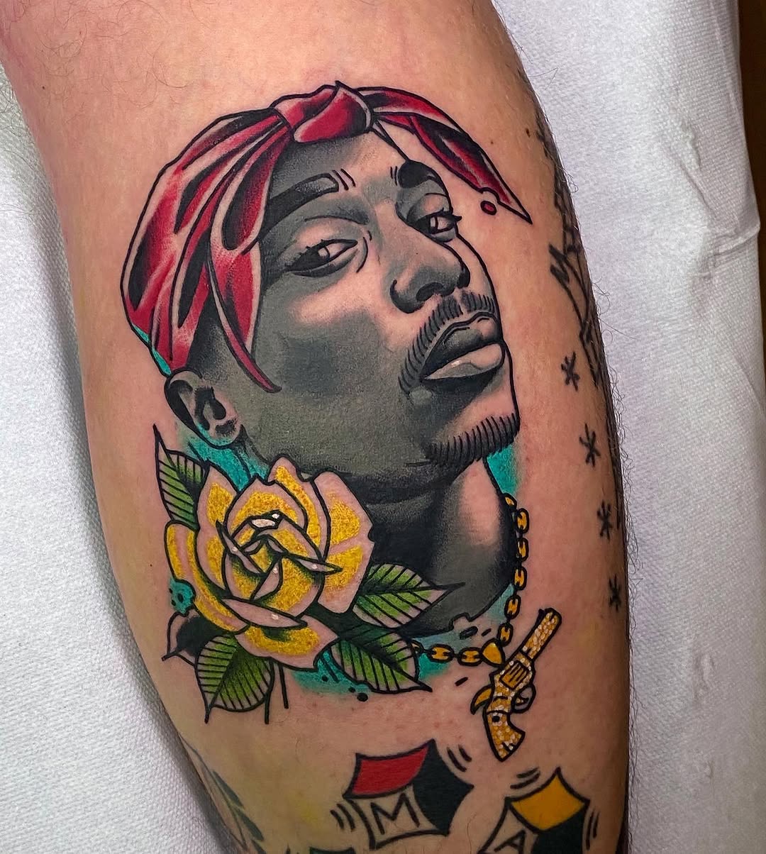 Colorful portrait tattoo on upper arm featuring a man with red hair, yellow rose, gold chain, and gun charm, with stars and geometric shapes below.