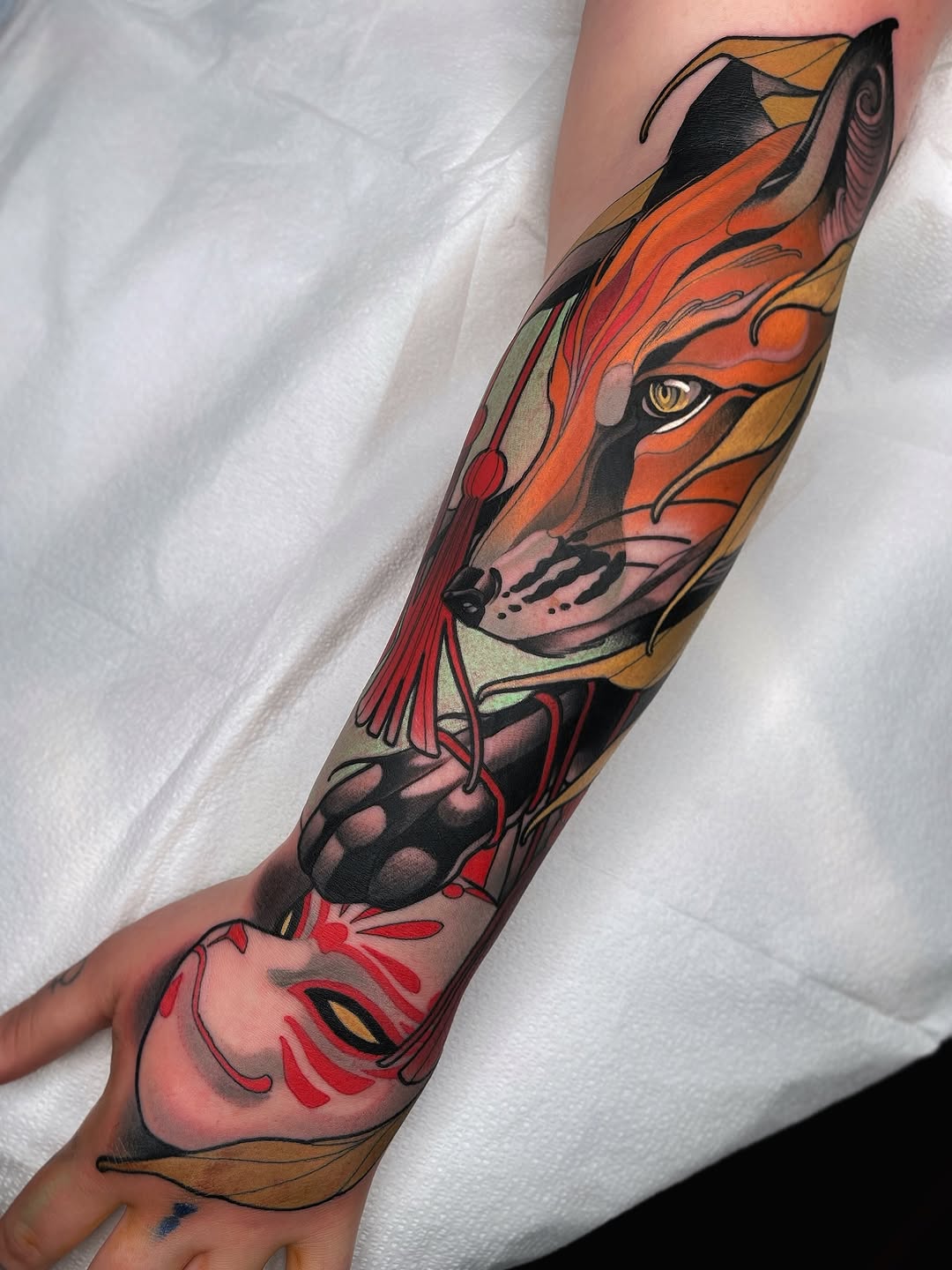 Colorful lion and fish head tattoo on forearm featuring orange, red, gold, and black with detailed facial features and flowing elements.