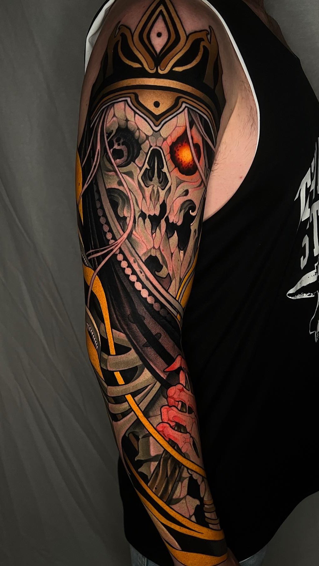 Skeletal skull with glowing red eyes and ornate crown on upper arm, featuring gold and black geometric patterns wrapping down the sleeve.