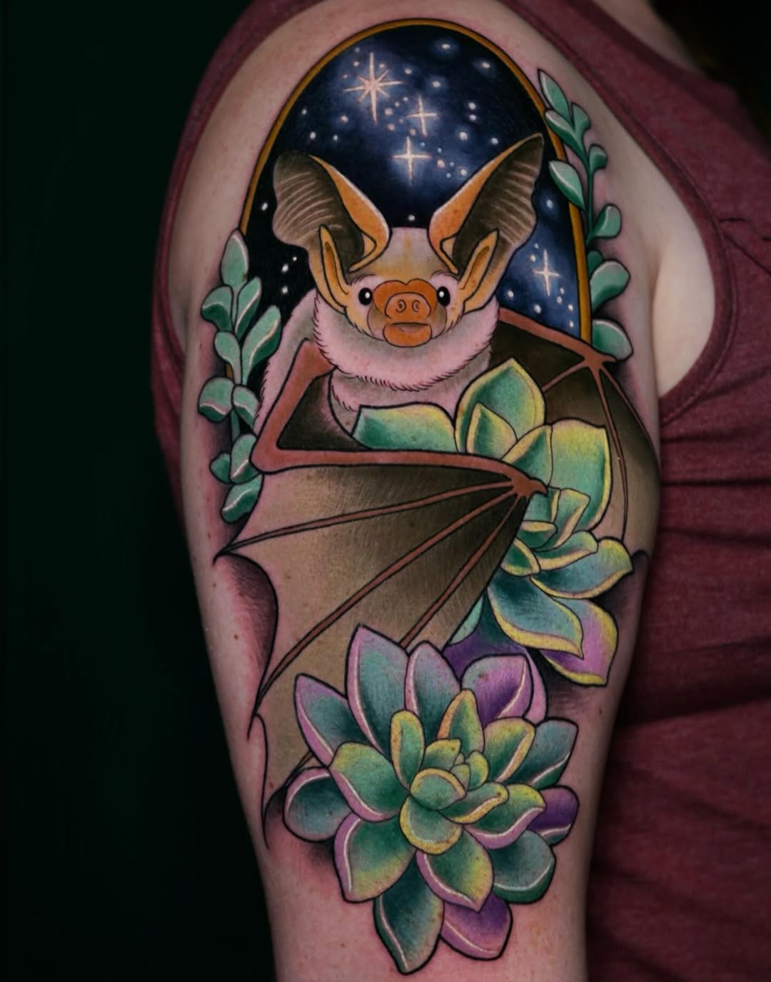 Colorful bat with pink fur and large ears surrounded by succulents and flowering plants against a starry night sky, tattooed on upper arm.
