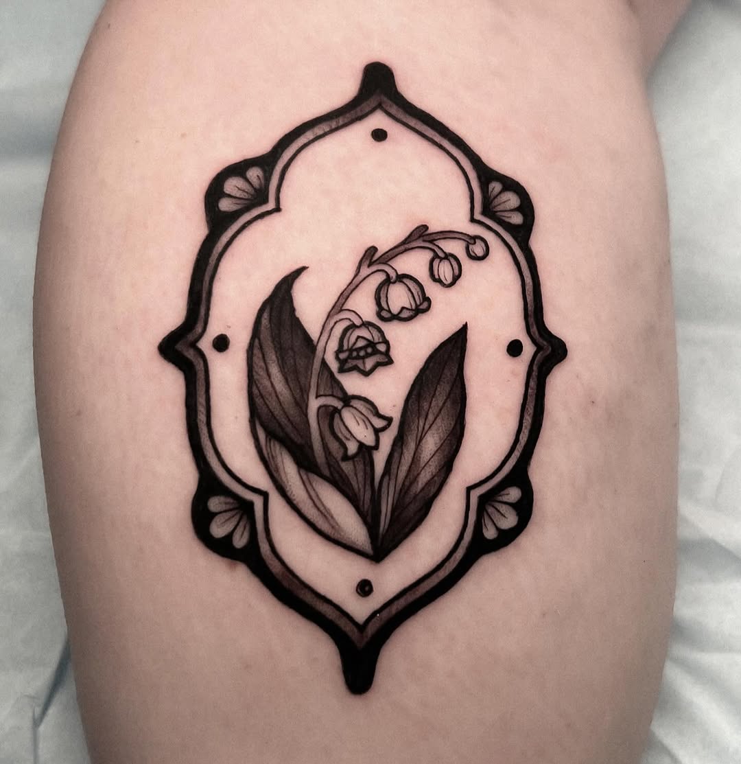 Lily of the valley flowers with leaves in an ornate oval frame with decorative corner elements, tattooed on the thigh in black ink.