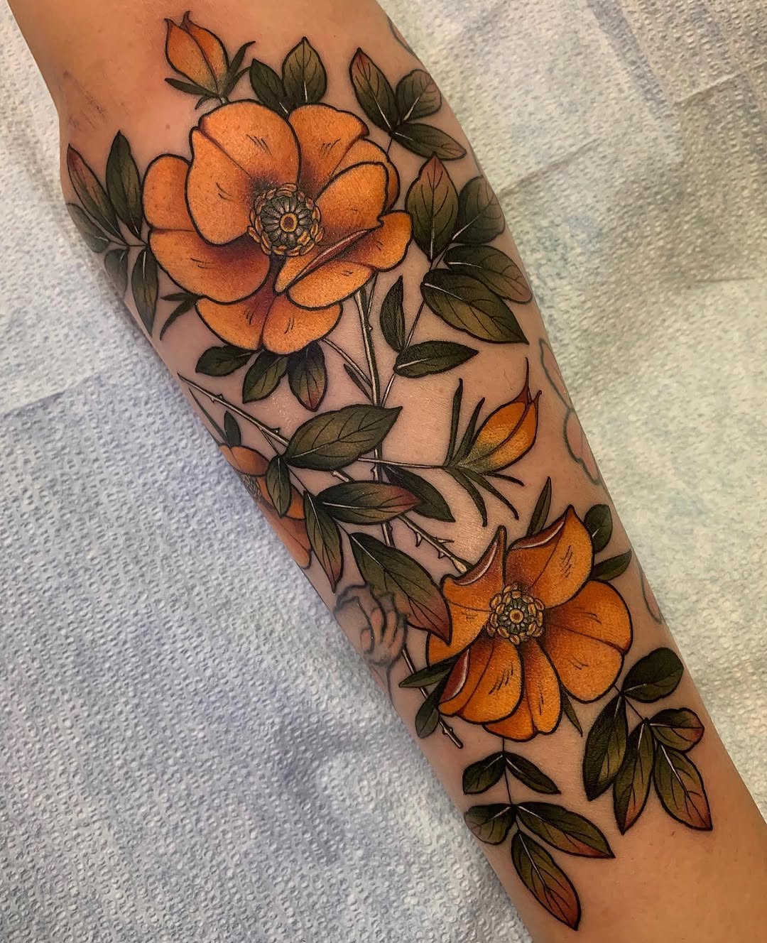Orange flowers with detailed centers and olive-green leaves cascade down the forearm in a vertical arrangement on tan skin.