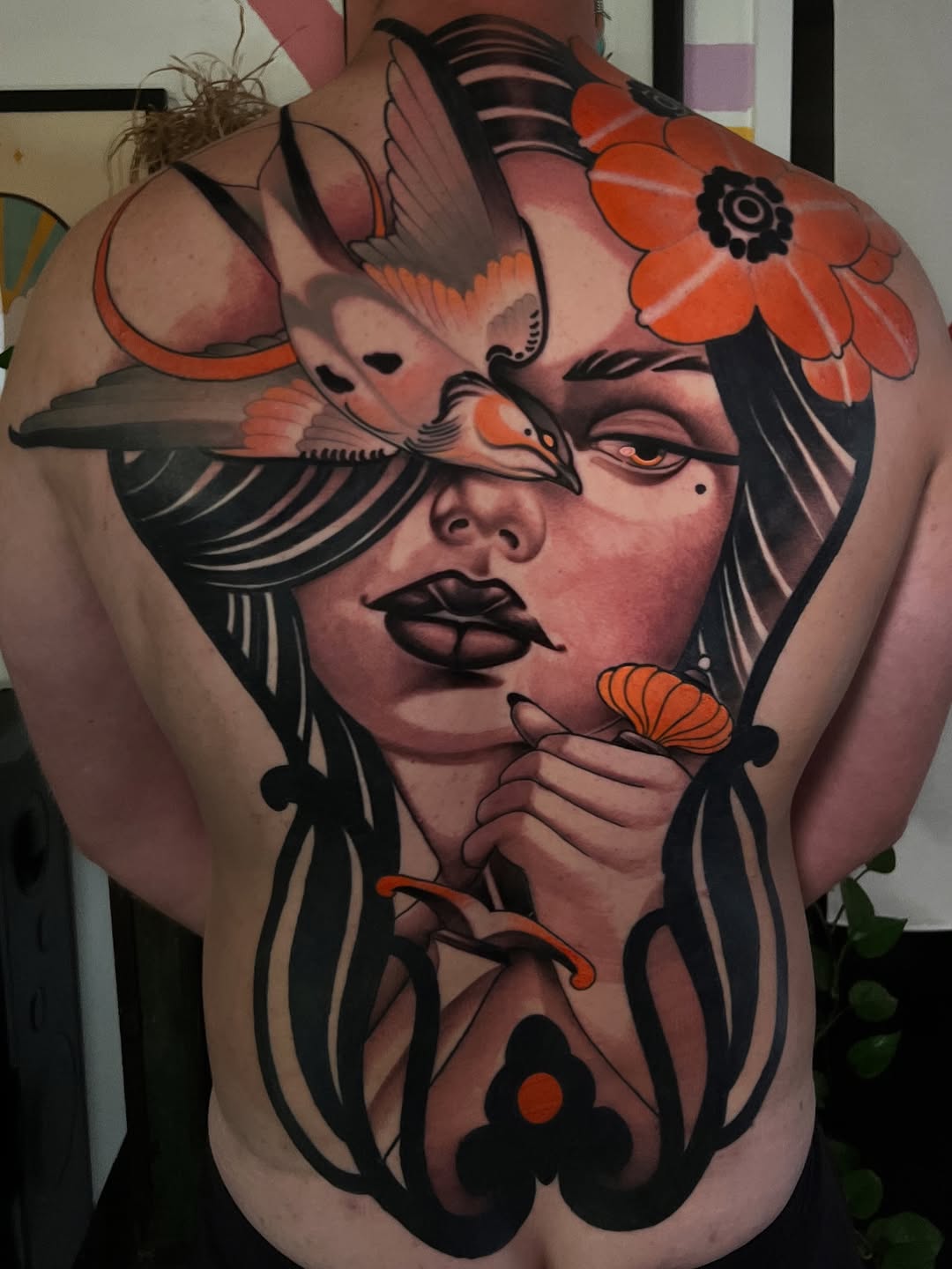 Large-scale back tattoo featuring a woman's face with birds and orange flowers, bold black linework and warm skin-tone shading throughout.