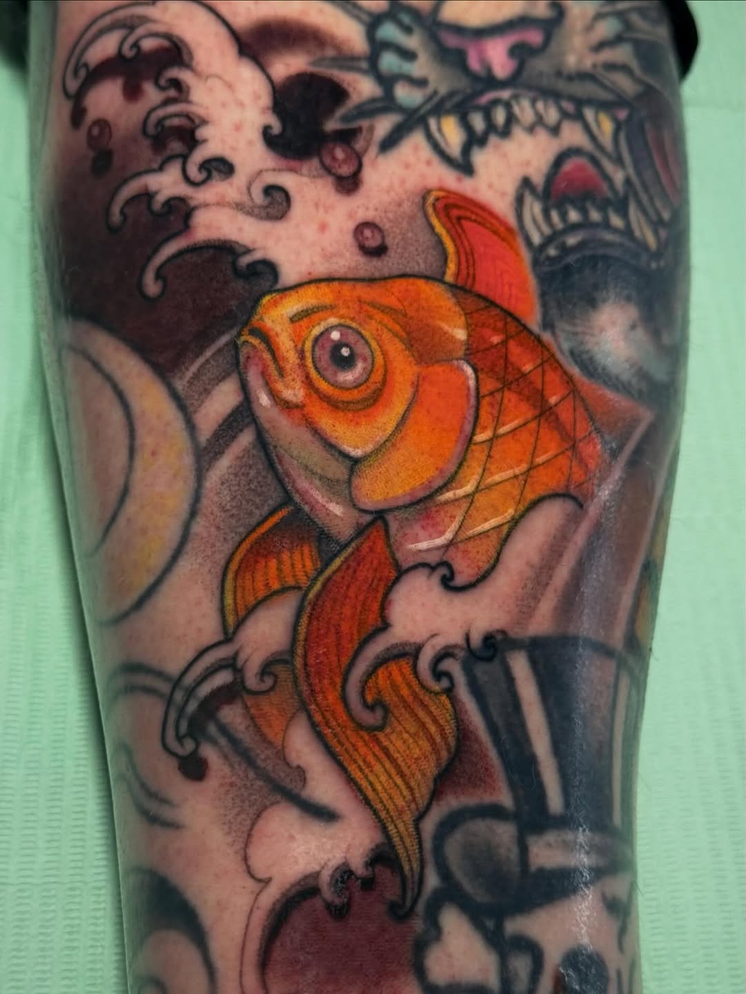 Colorful orange and red goldfish tattoo on upper arm with decorative black swirls and darker design elements in the background.