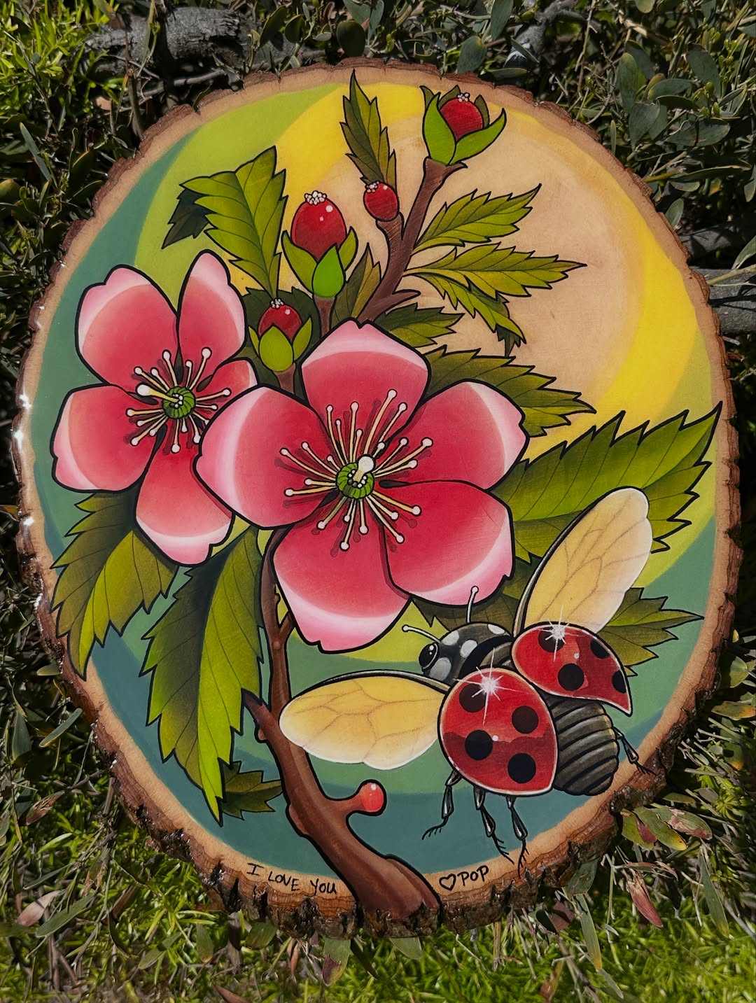 Colorful tattoo on a wood slice featuring pink flowers, red berries, green leaves, a ladybug, and text reading "I LOVE YOU" and "POP" on the natural wood surface.