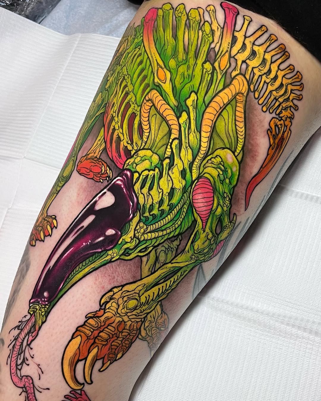 Colorful creature tattoo on calf featuring a green and yellow alien-like head with tentacles, red mouth, and orange clawed feet against tan skin.
