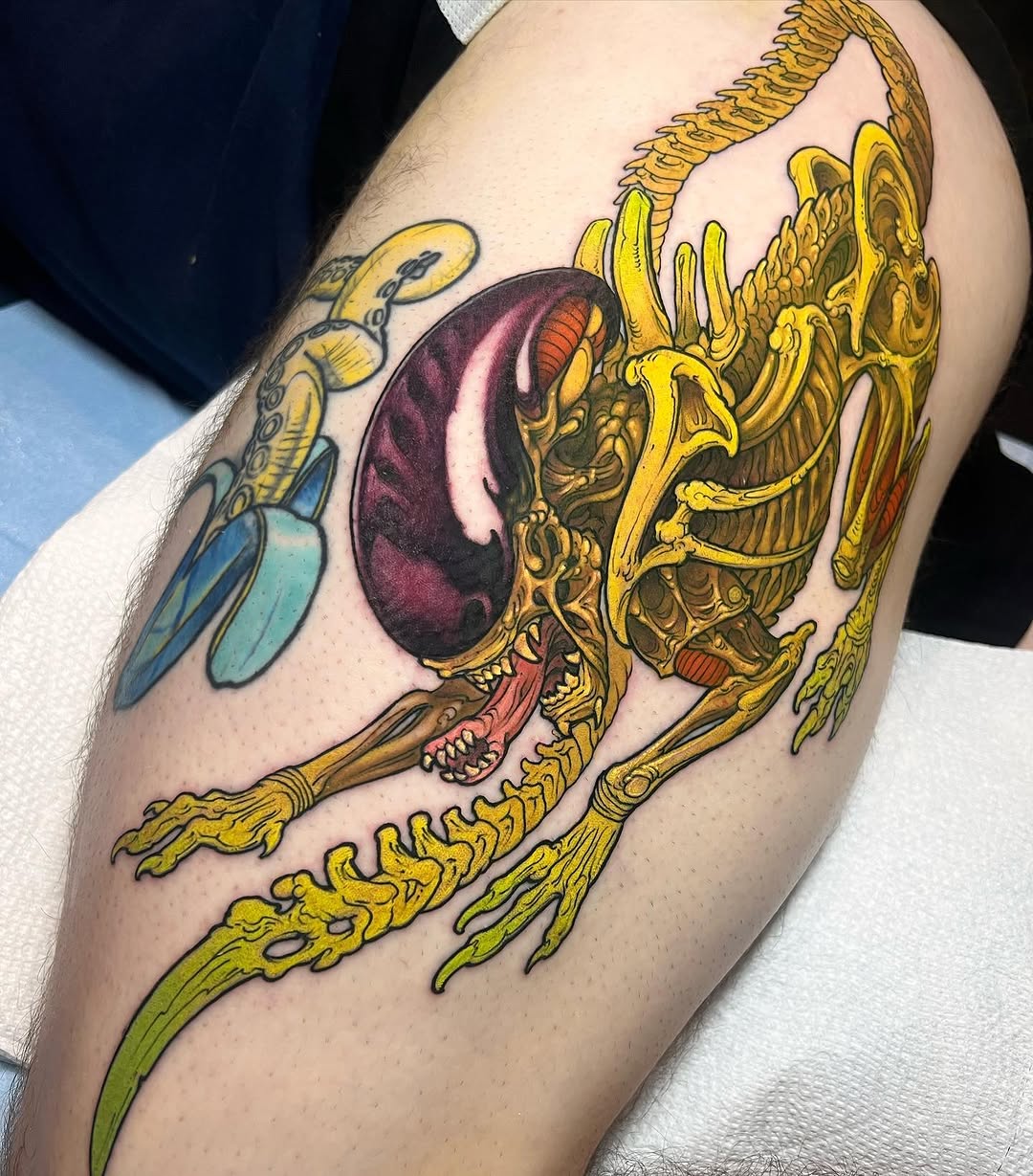 Colorful anatomical eye tattoo with golden ornamental wings and claws on upper arm, featuring purple, yellow, and green tones.