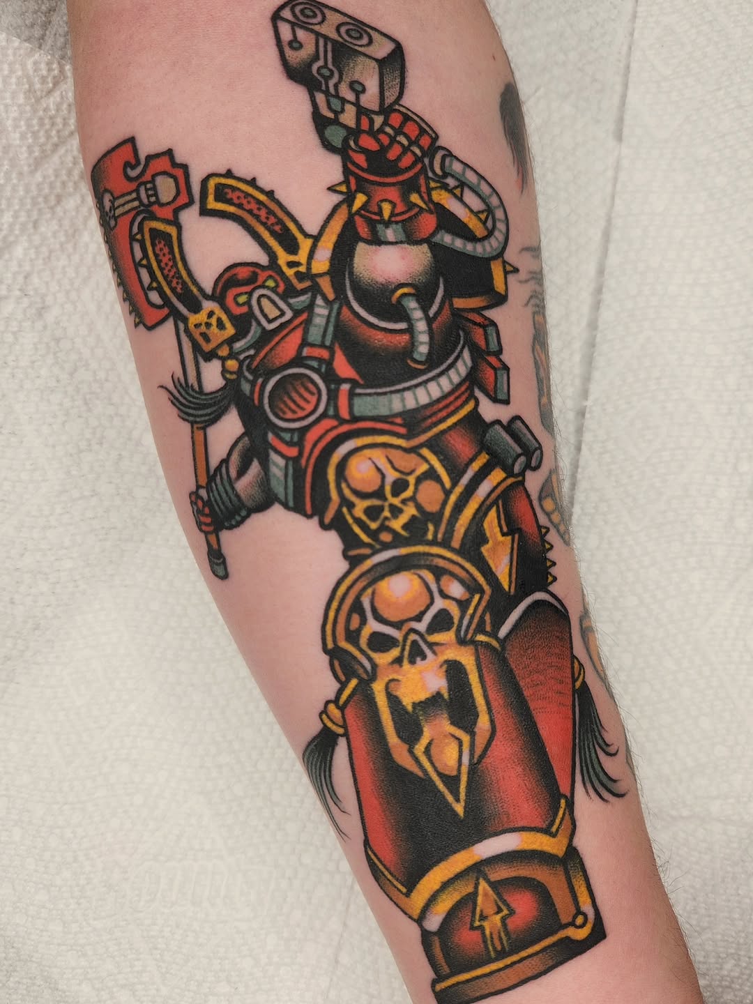 Colorful robotic figure tattoo on forearm featuring red, orange, and black mechanical body with geometric skull designs and weapon details.