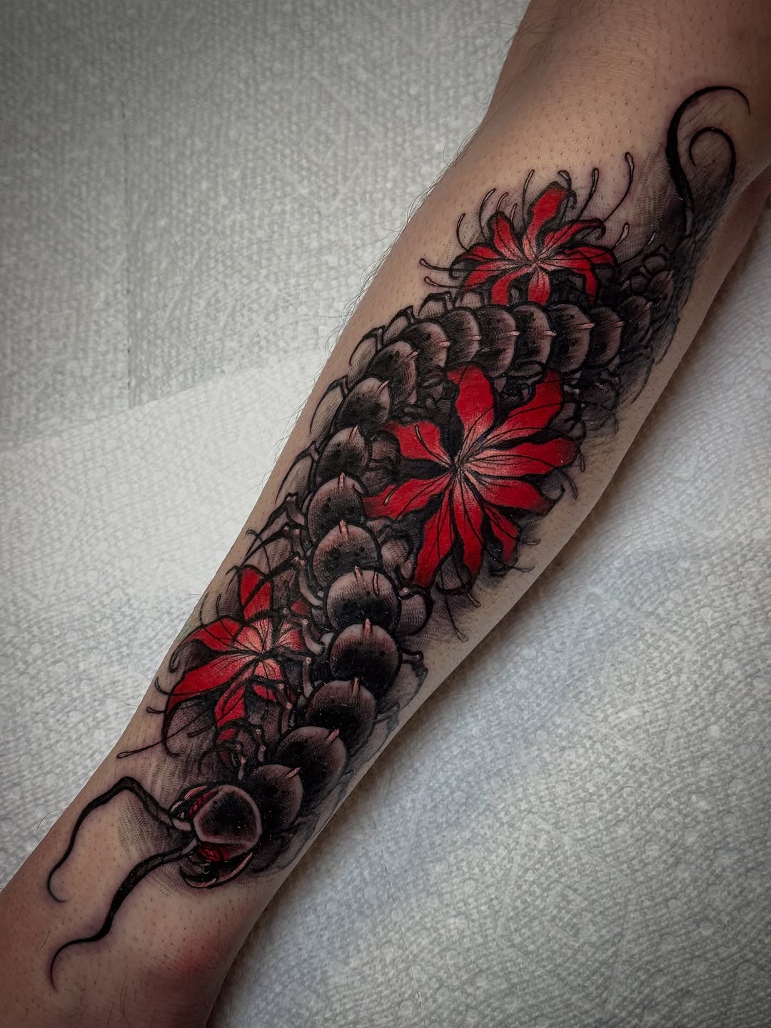 Vertical forearm tattoo featuring red flowers with dark botanical elements and curved vine details extending from wrist to elbow.