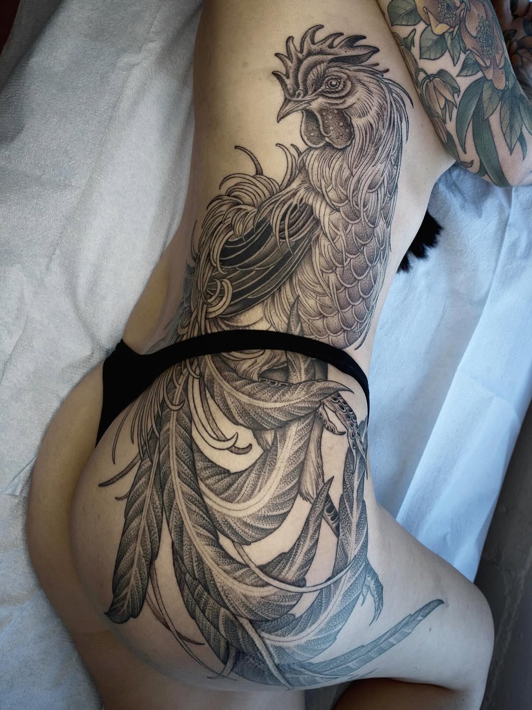 Detailed rooster head and flowing feather tattoo on thigh in black and grey ink with intricate line work and shading.