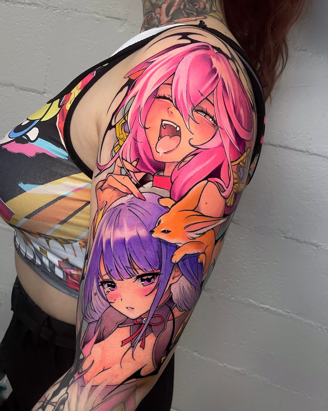 Colorful sleeve tattoo featuring two anime characters with pink and purple hair, accompanied by an orange fox, displayed on an arm against a brick wall.