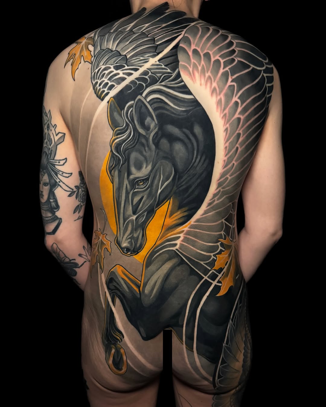 Large-scale winged horse tattoo spanning the back in black, grey, and gold with feathered wings extending across the shoulders and sides.