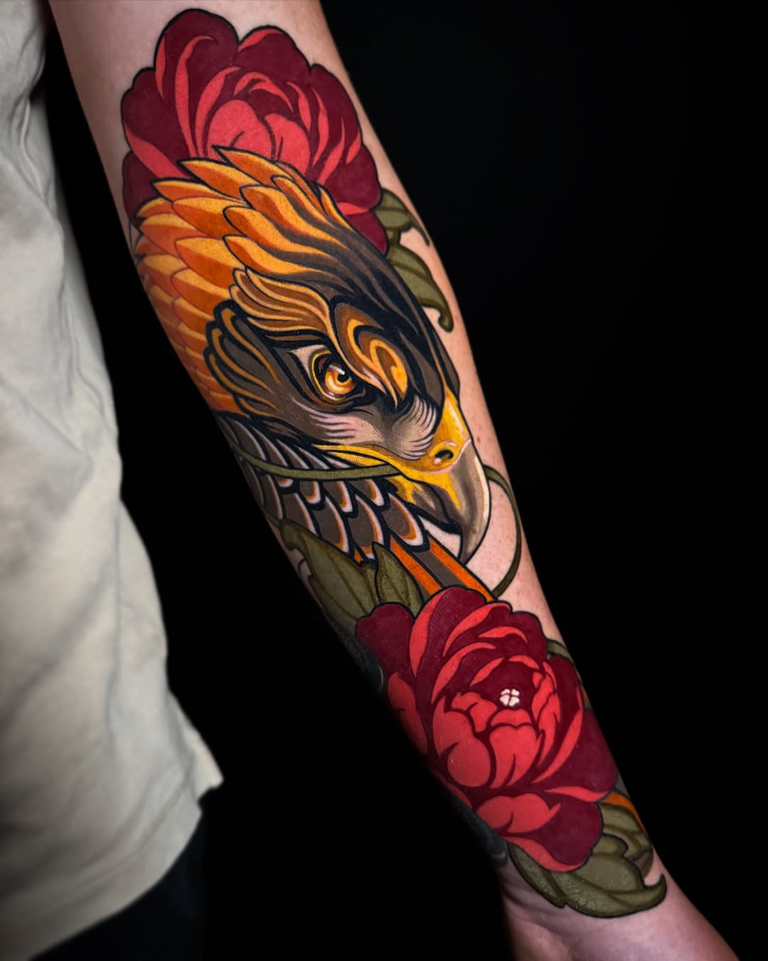 Eagle head with yellow and orange plumage surrounded by deep red roses with green leaves on a forearm tattoo.