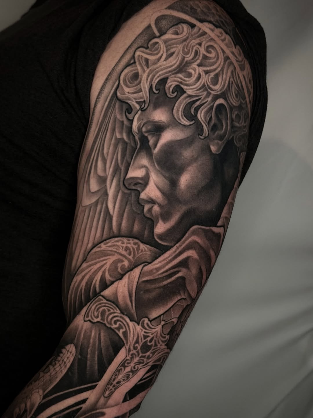 Detailed portrait tattoo on upper arm depicting two male faces in profile with ornate curled hair and draped fabric, rendered in grayscale.