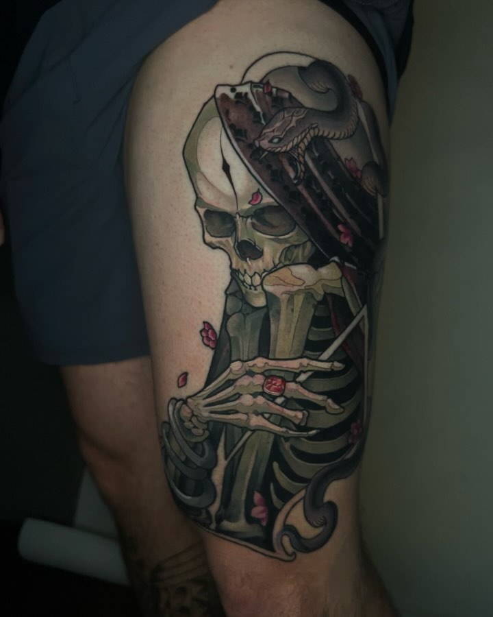 Skull with skeletal hands holding roses and a raven perched on top, rendered in black, green, and red ink on the upper thigh.